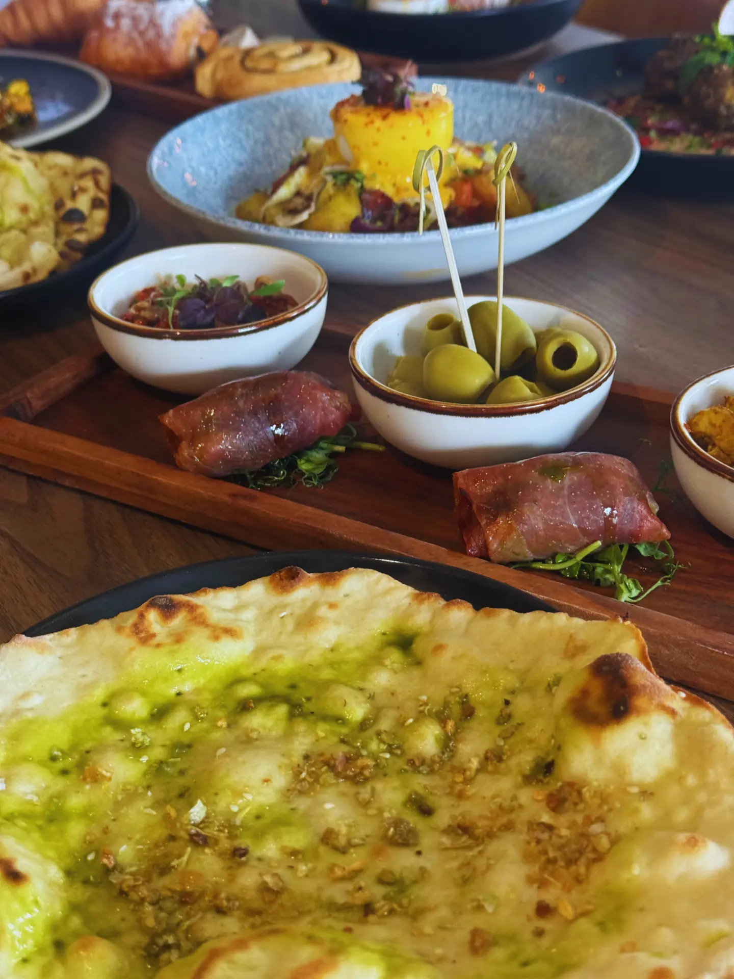 Warm stone-baked garlic and za'atar flatbread with herb oil, with olives and prosciutto parcels on the table behind